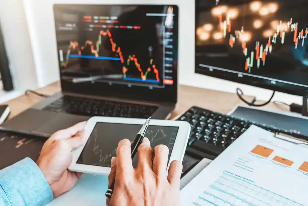 Person analyzing financial data on a tablet, surrounded by monitors displaying stock market charts and a keyboard, with papers featuring graphs and numbers on a desk.