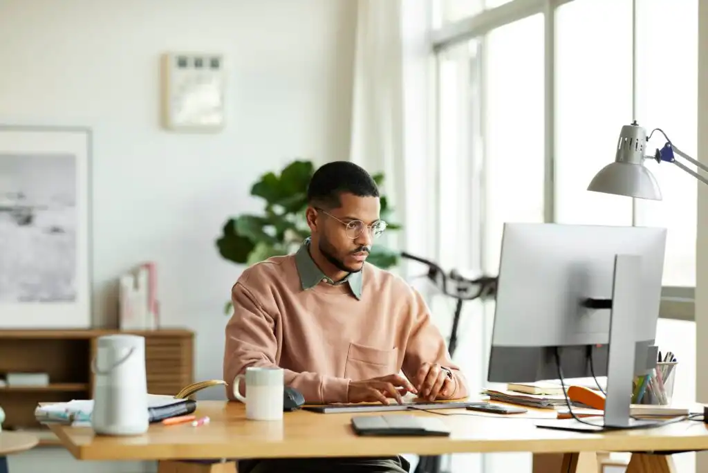 A man wearing glasses and a peach sweater sits at a desk, working on a computer in a bright, modern office with large windows, plants, and various office supplies around him.