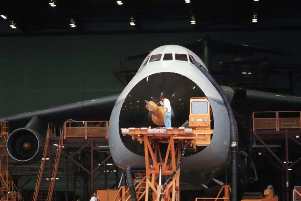 A worker stands on a platform inside the open nose of a large cargo aircraft in a hangar, surrounded by scaffolding and equipment.