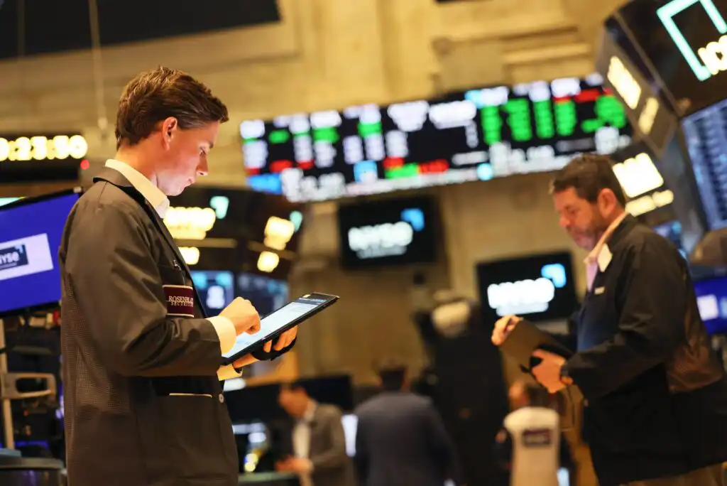 Two people in professional attire use tablets on a busy stock exchange floor, with digital stock tickers and financial data displayed on large screens in the background.