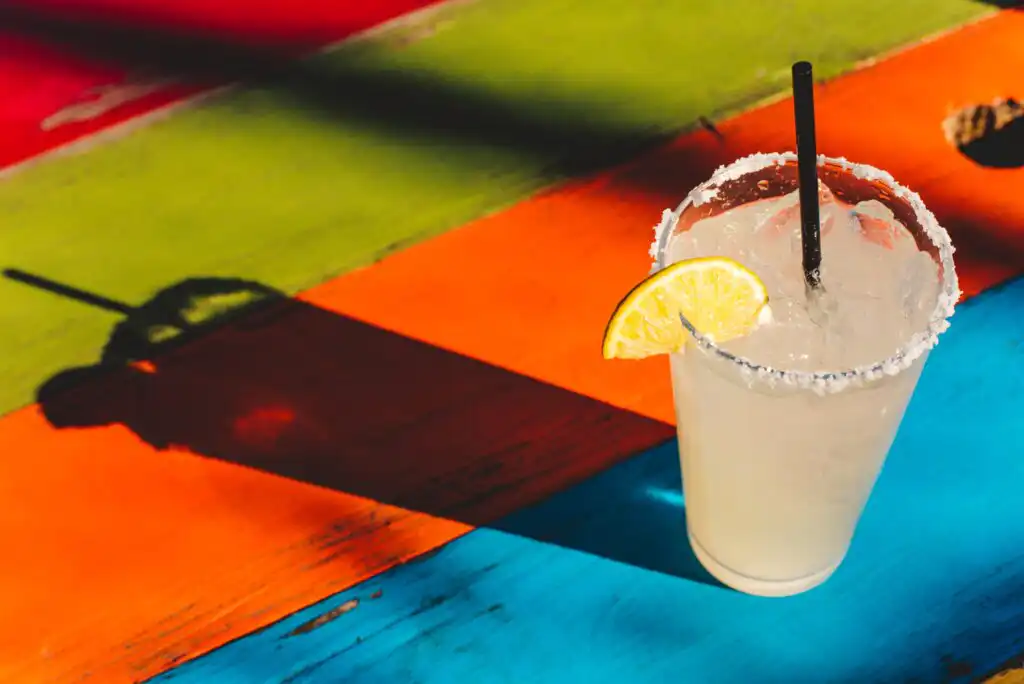 A margarita with a salted rim, lime wedge, and straw sits on a colorful wooden table with orange, green, blue, and red sections, casting a shadow in bright sunlight.