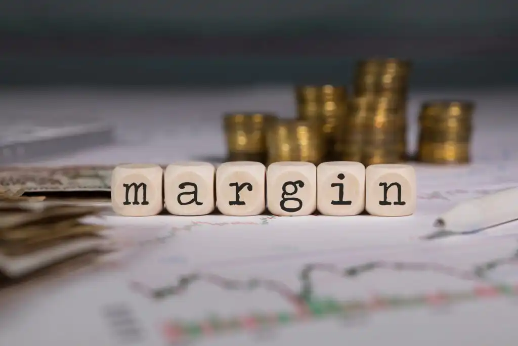 Wooden blocks spelling margin are placed on financial charts, with stacks of coins and a pen in the blurred background, representing financial or investment concepts.