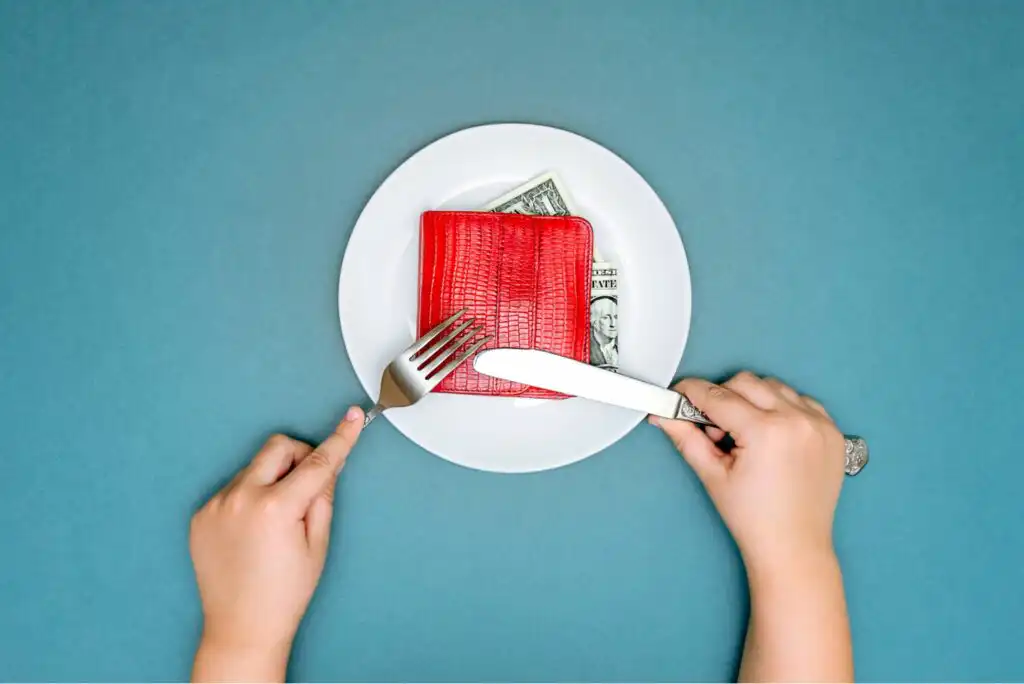 A persons hands hold a fork and knife over a white plate containing a red wallet and dollar bills, set against a blue background.