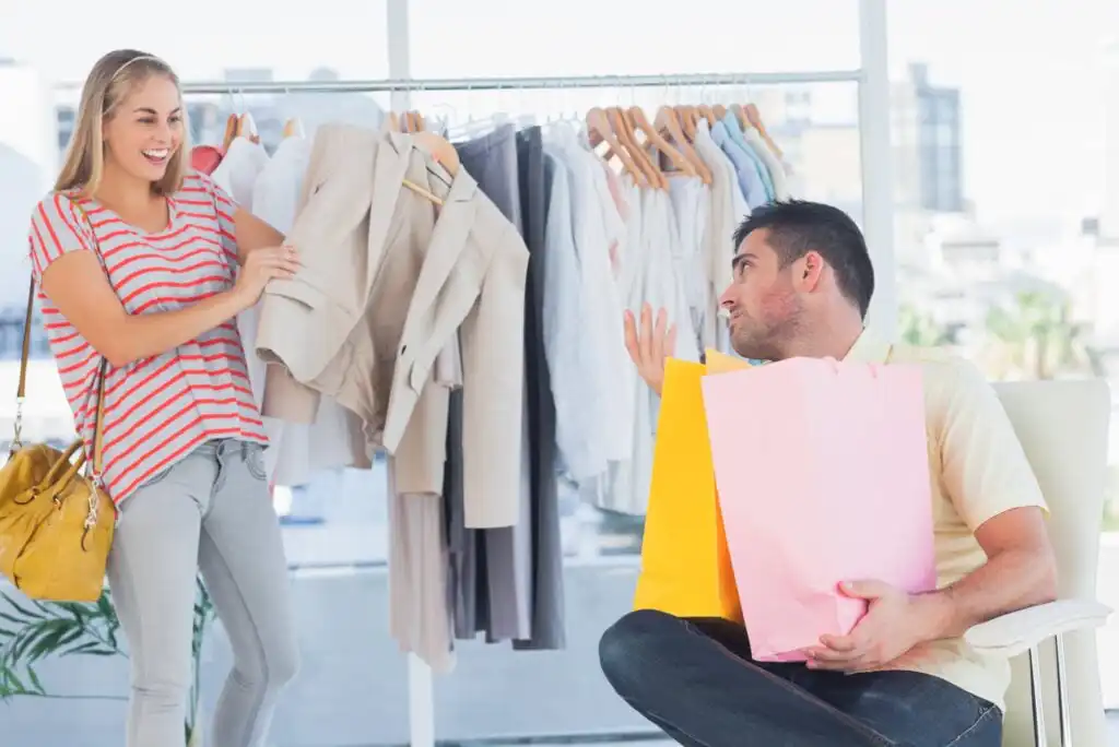 A smiling woman shows clothes on hangers to a seated man holding shopping bags in a bright clothing store.