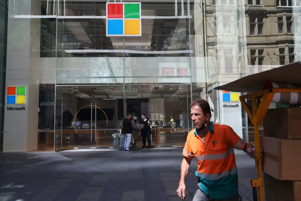 A man in an orange high-visibility shirt pushes a cart in front of a Microsoft store with large glass windows and the Microsoft logo displayed above the entrance. Several people are visible inside the store.