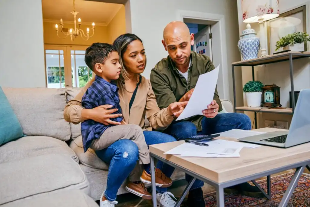 A couple sits on a couch with their young son, reviewing papers together at a coffee table with a laptop, calculator, and documents, appearing focused and concerned in a home setting.