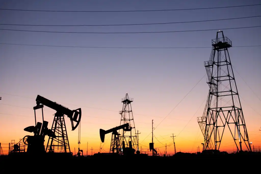 Silhouettes of oil pumpjacks and drilling rigs stand against a colorful sunset sky, with power lines running overhead.