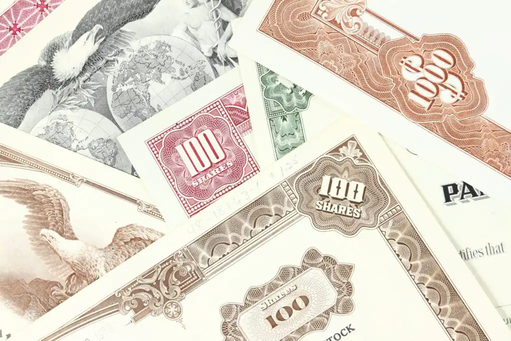 A close-up of several vintage stock certificates in different colors, featuring ornate borders, the word “shares,” and illustrations of eagles and globes.