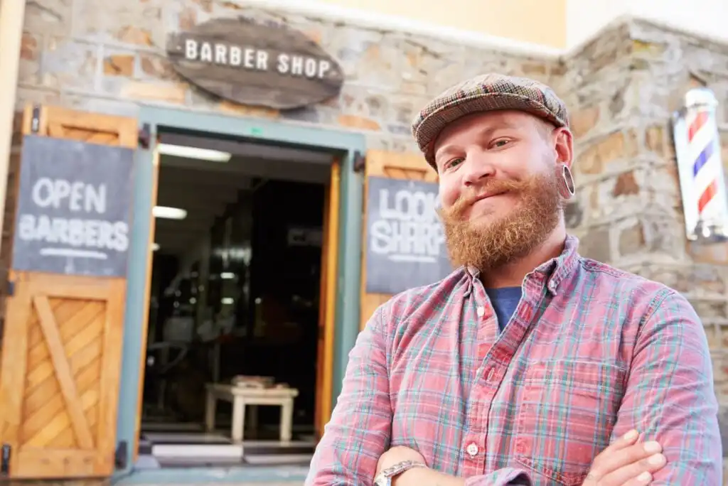 A bearded man with a plaid shirt and flat cap smiles confidently with arms crossed in front of a rustic barbershop, with signs reading OPEN BARBERS and LOOK SHARP visible near the entrance.