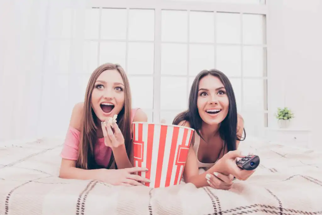 Two young women lying on a bed, smiling, watching TV. One is holding a remote, the other eating popcorn from a large striped bucket. Bright window and plaid blanket in the background.