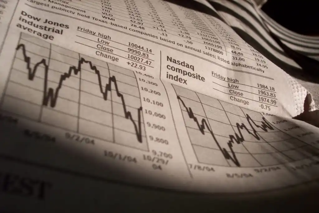 A close-up of a newspaper showing stock market charts and data tables for the Dow Jones Industrial Average and Nasdaq Composite Index, with fluctuating line graphs and numerical values under a shadow.