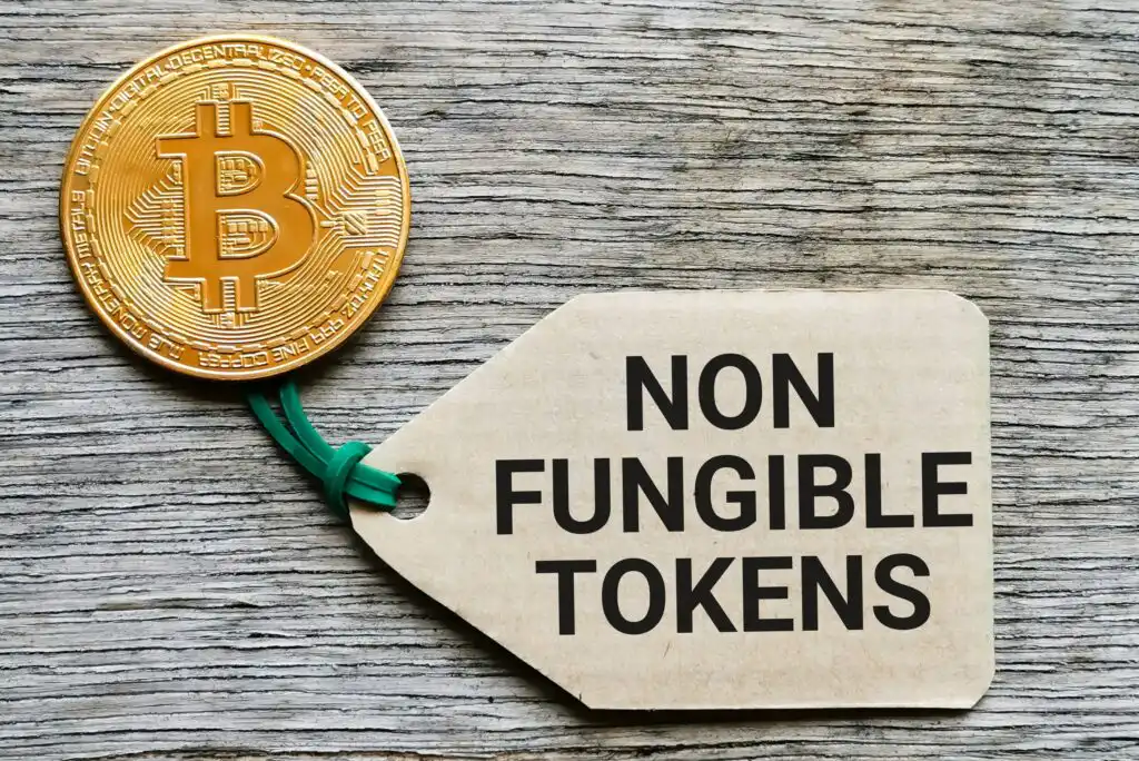 A gold Bitcoin token is placed next to a brown paper tag labeled NON FUNGIBLE TOKENS on a wooden surface. The tag is attached with a green string.