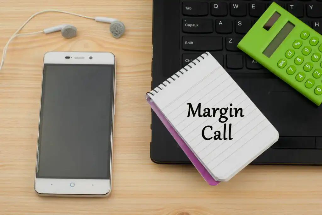 A notepad with Margin Call written on it lies on a desk next to a smartphone, a green calculator, earphones, and a laptop keyboard.