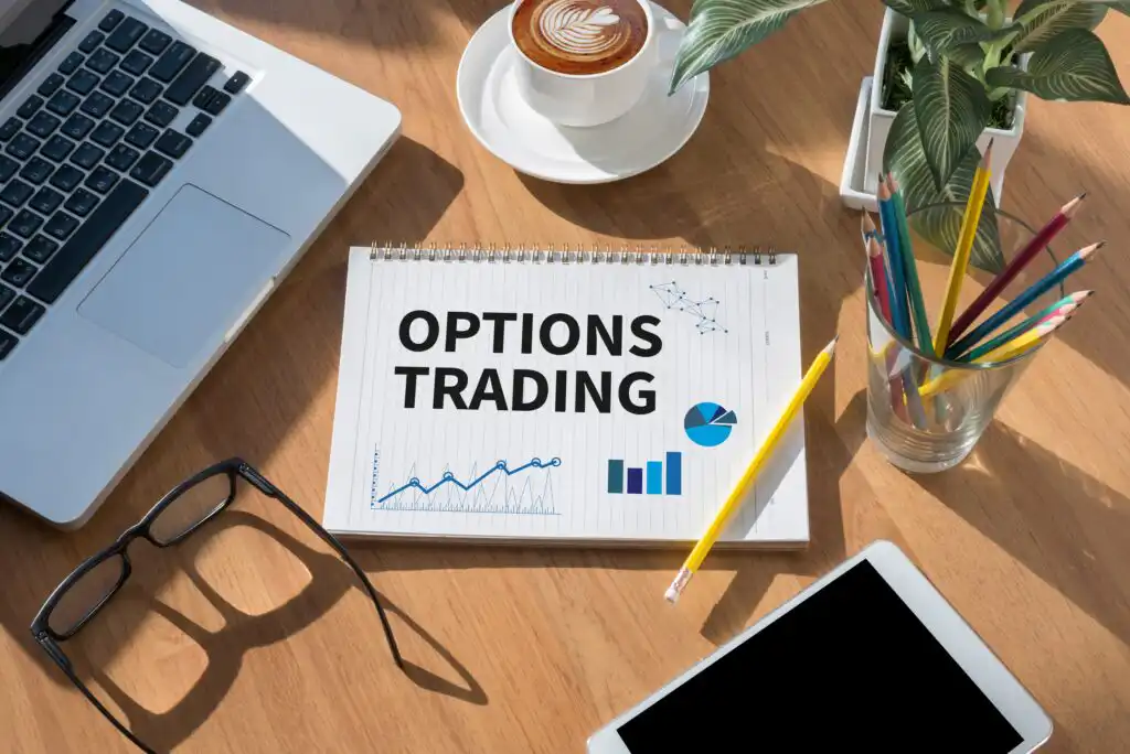 A notepad with OPTIONS TRADING and charts on it sits on a desk with a laptop, a cup of coffee, a plant, colored pencils in a glass, and a tablet.