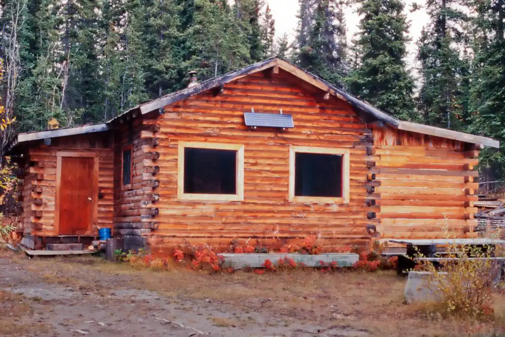A wooden log cabin with a solar panel above the front windows sits in a forest clearing, surrounded by trees and autumn foliage, with a bench and table nearby.