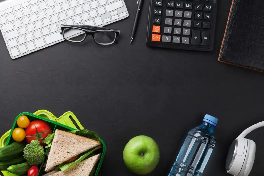 A workspace with a keyboard, glasses, pen, calculator, notebook, headphones, bottled water, green apple, and a lunchbox with sandwiches and vegetables on a dark desk.