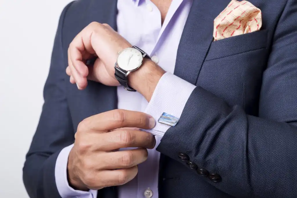 A person in a dark blue suit jacket adjusts their shirt cuff, showing a wristwatch and cufflink. The shirt is light purple, and a patterned pocket square is visible in the jacket pocket.