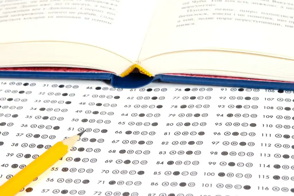 A sharpened yellow pencil rests on a standardized multiple-choice answer sheet, with an open book lying in the background. The answer bubbles are filled in, indicating the test is in progress or completed.
