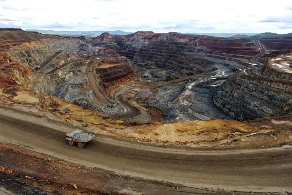A large open-pit mine with terraced layers of exposed earth and rock. A dump truck loaded with ore drives along a dirt road in the foreground. The landscape is rugged with varying shades of brown and red.