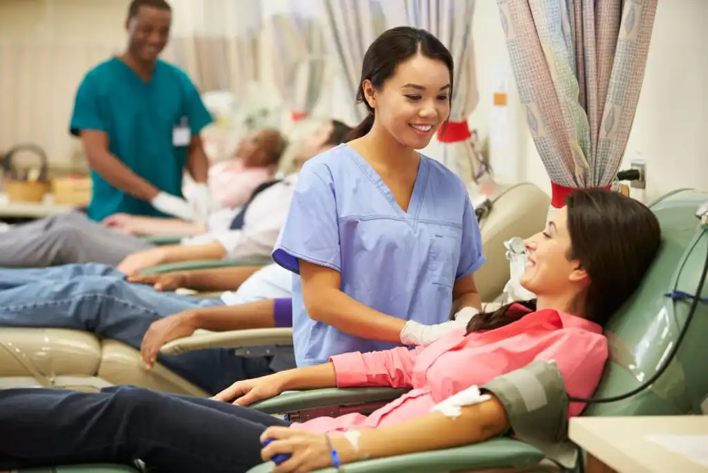 A nurse smiles and speaks to a woman donating blood in a clinic. Other donors are seated in the background, and a staff member stands near them. The setting appears clean and friendly.