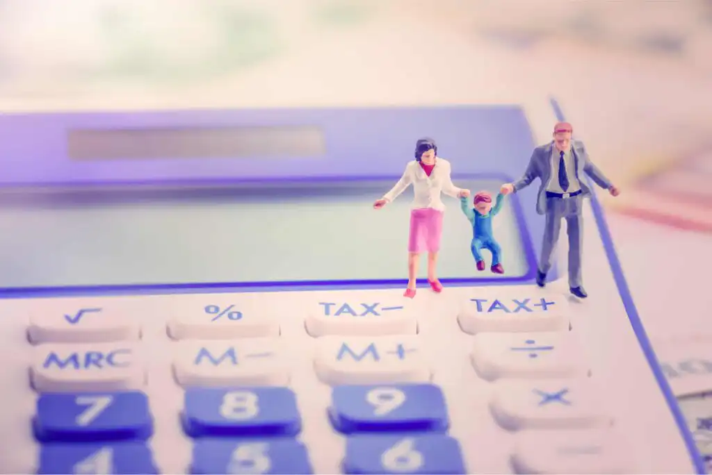 Miniature figurines of a family—mother, father, and child—stand on a calculator. The calculator keys are labeled with tax-related words like TAX- and TAX+. The scene represents finances and taxation for families.