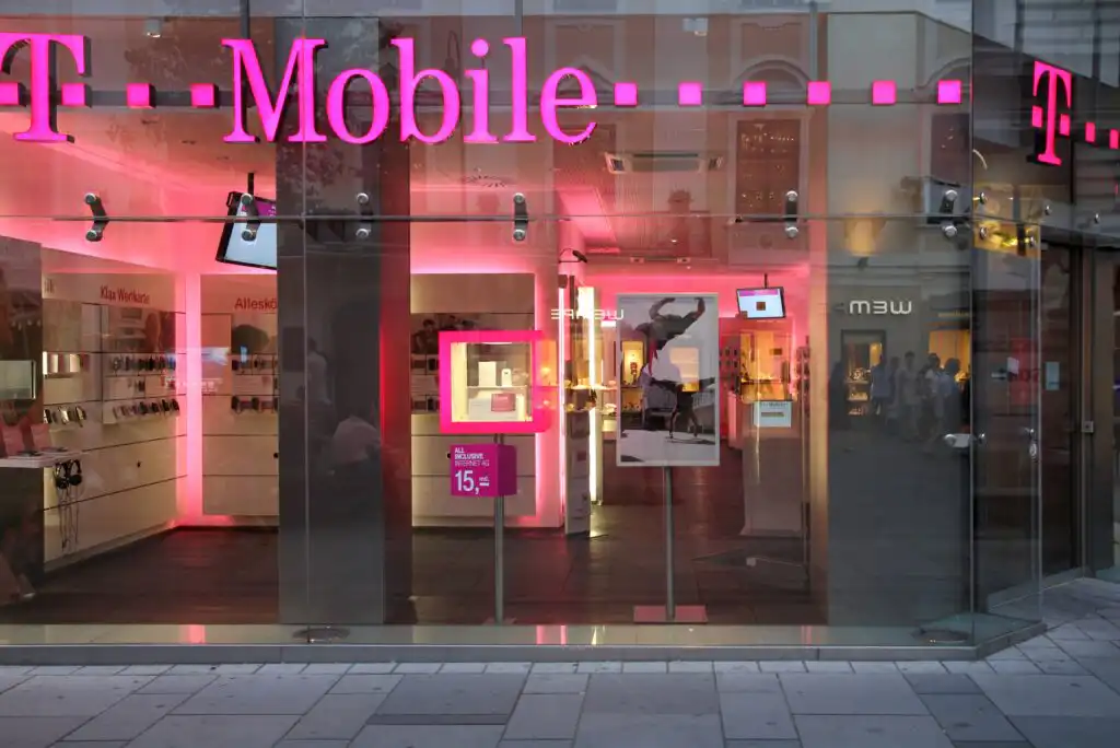 A T-Mobile store with glass walls and bright pink lighting. Inside, displays of phones and accessories are visible, along with product posters and a digital sign showing a promotion.