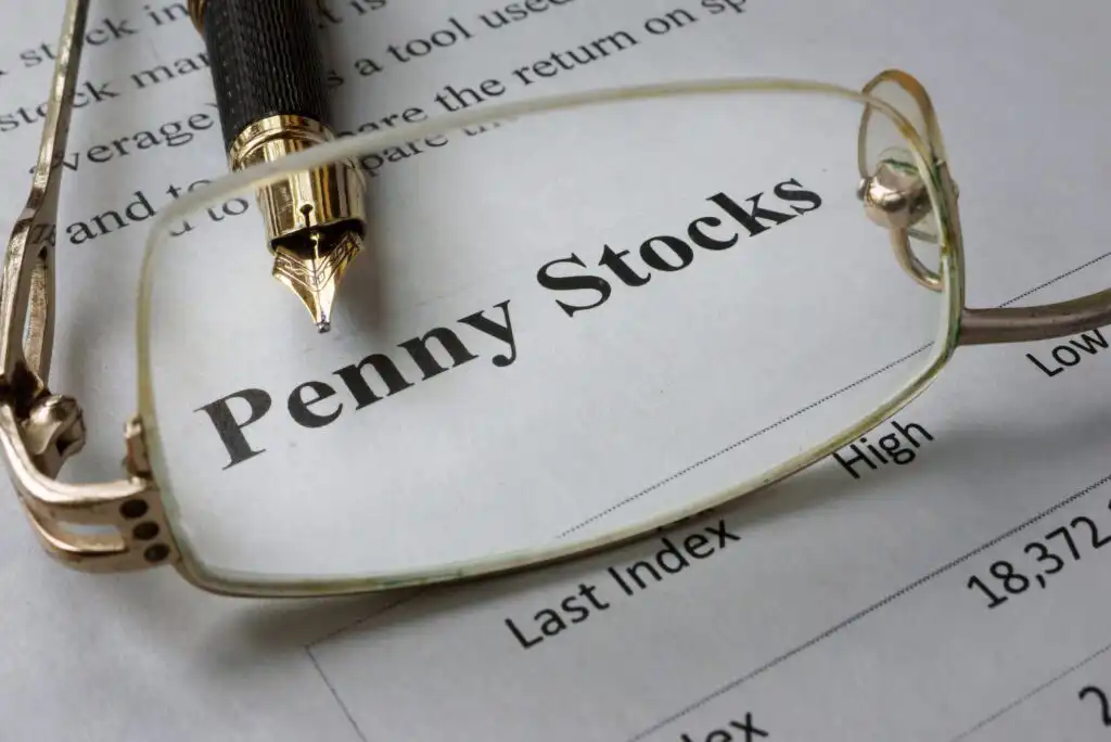 A close-up of eyeglasses and a fountain pen resting on a document with the bold heading Penny Stocks. The text is in focus through the glasses, suggesting analysis or review.