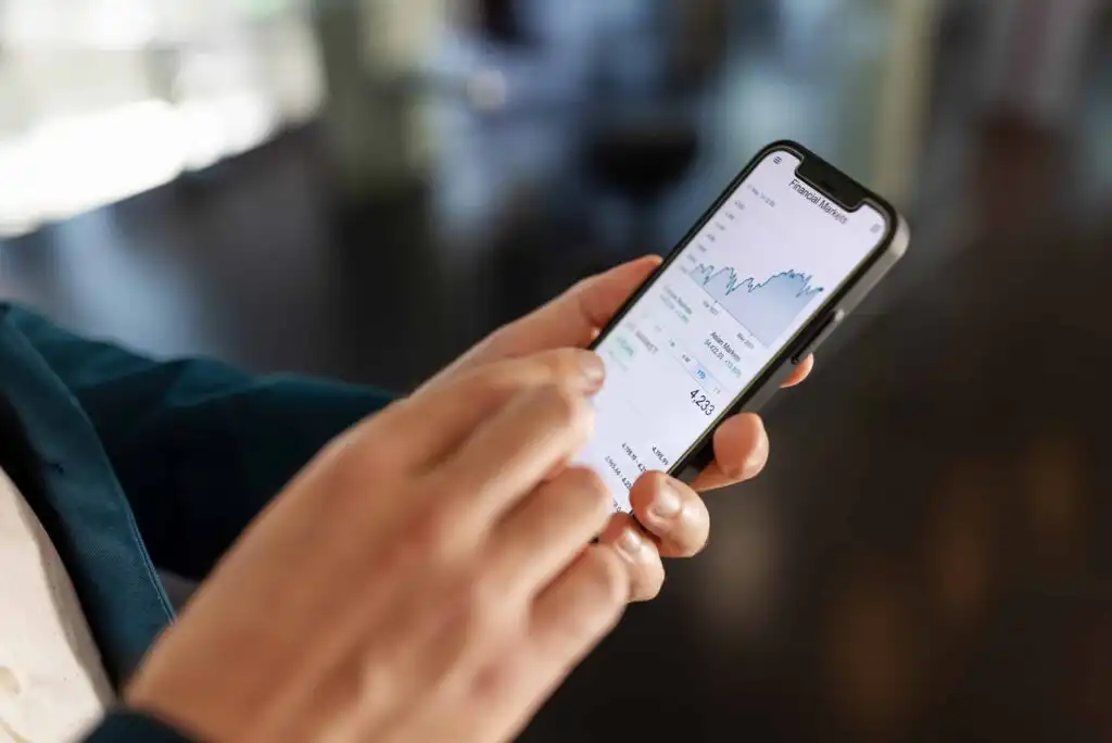 A person holds a smartphone displaying a financial app with a line graph and market data, suggesting they are monitoring or trading stocks. The background is blurred.