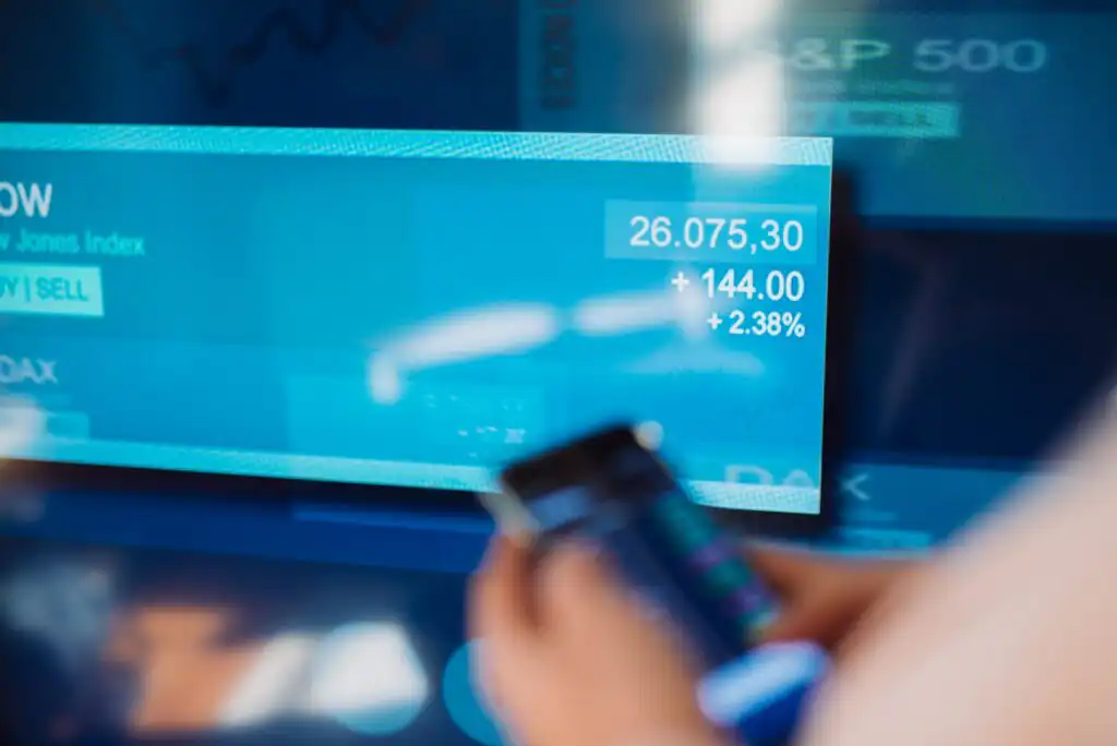 A blurred hand holds a smartphone in front of a digital screen displaying stock market data showing an increase of 144.00 points and a 2.38% rise, with blue and white financial graphics in the background.
