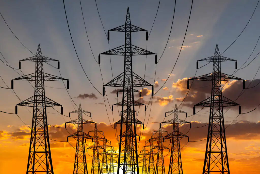 Multiple high-voltage power lines and transmission towers stretch into the distance against a dramatic orange and yellow sunset sky, with clouds scattered across the horizon.