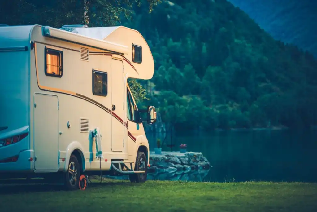 A white camper van is parked on grass near a lake, with forested hills in the background at dusk. Warm lights glow from the camper’s window, creating a cozy atmosphere by the tranquil water.
