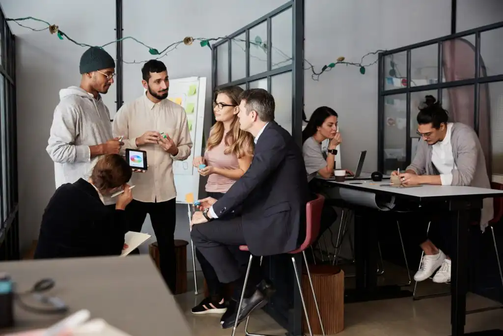 A group of people collaborates in a modern office. Five individuals stand and sit in discussion near a glass wall, while two others work separately at desks. Laptops, notes, and string lights are visible.
