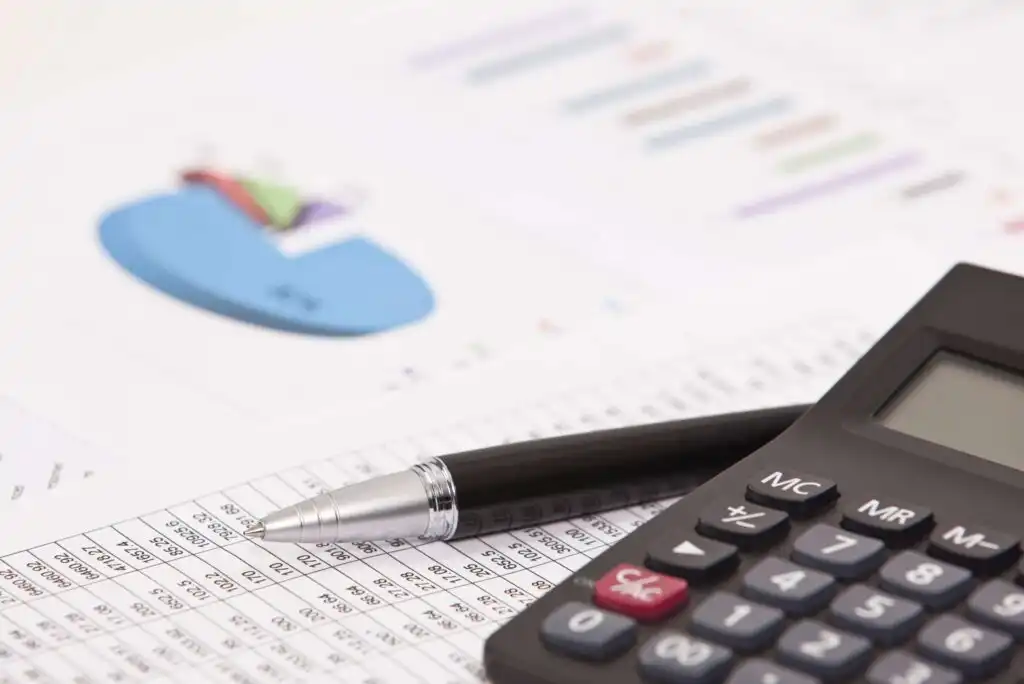 A black pen and a calculator rest on a financial spreadsheet, with a blurred pie chart and bar graphs visible in the background, suggesting financial analysis or accounting work.