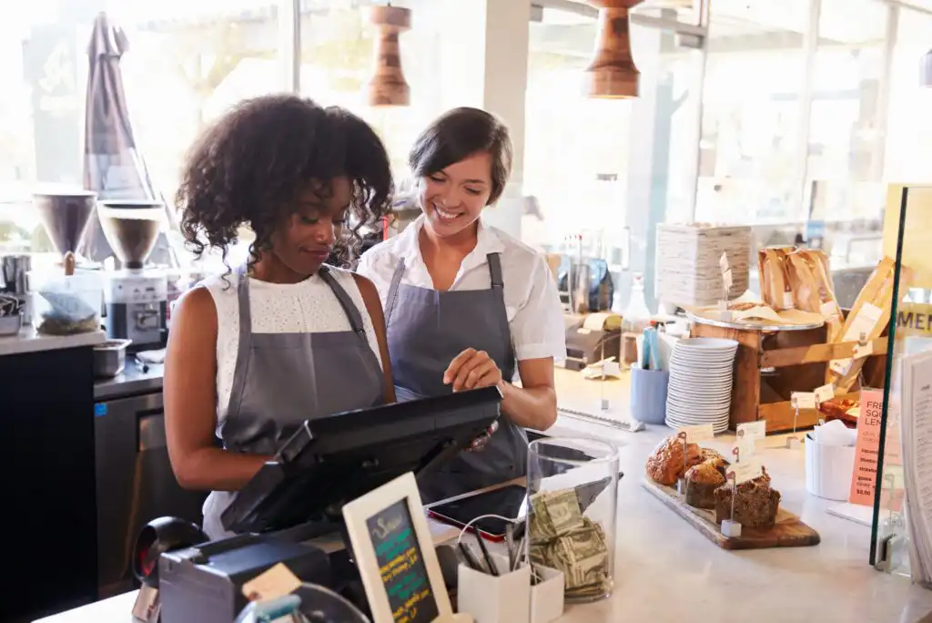 Two women wearing aprons stand behind a café counter; one operates the cash register while the other smiles and watches. Baked goods and a tip jar are visible on the counter.