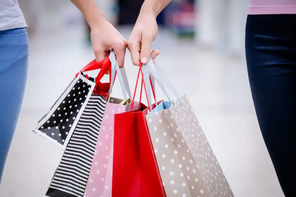 Two people hold several colorful shopping bags with different patterns, including polka dots and stripes, standing side by side in a brightly lit indoor setting.