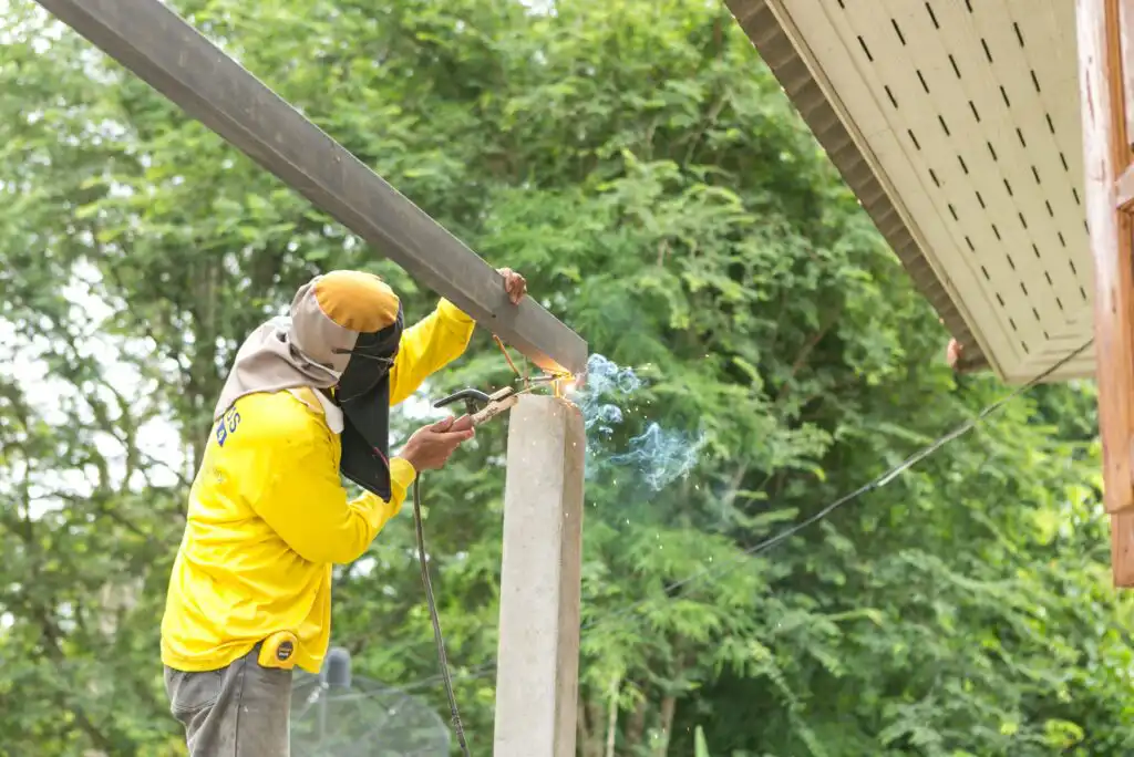 A person wearing a yellow shirt and protective head covering is welding a metal beam onto a concrete post outdoors, with trees and part of a roof visible in the background.