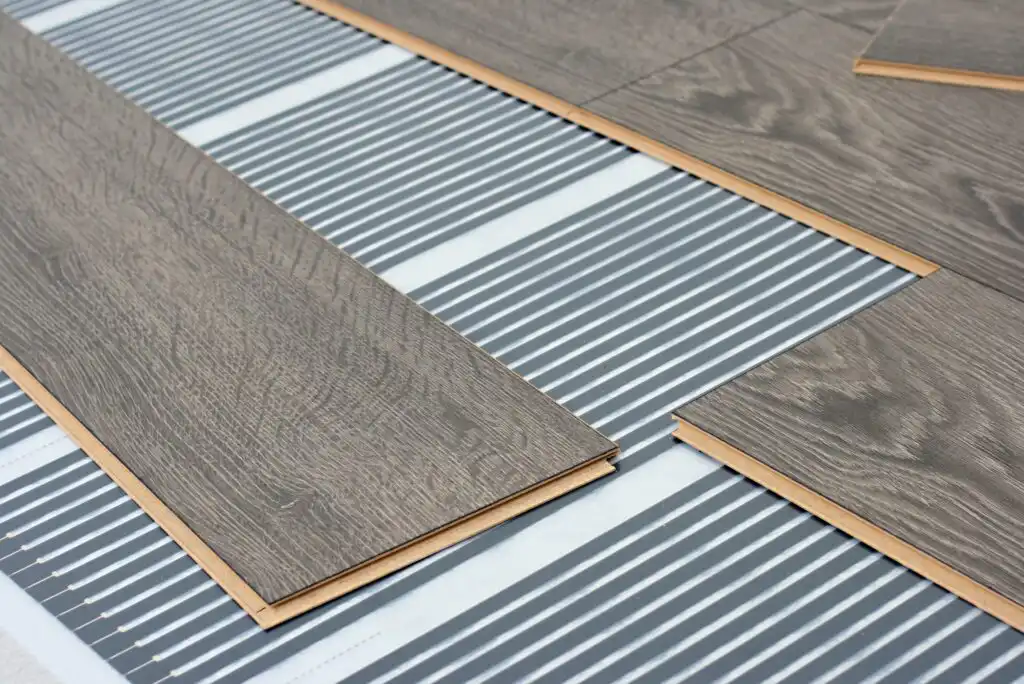 Close-up of wood laminate flooring panels being installed over an underfloor heating system with gray and white striped heating elements. The panels are partially assembled on top of the heating mat.