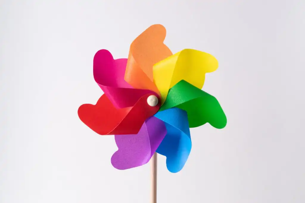 A colorful pinwheel with heart-shaped blades in red, orange, yellow, green, blue, and purple, attached to a wooden stick, set against a plain white background.