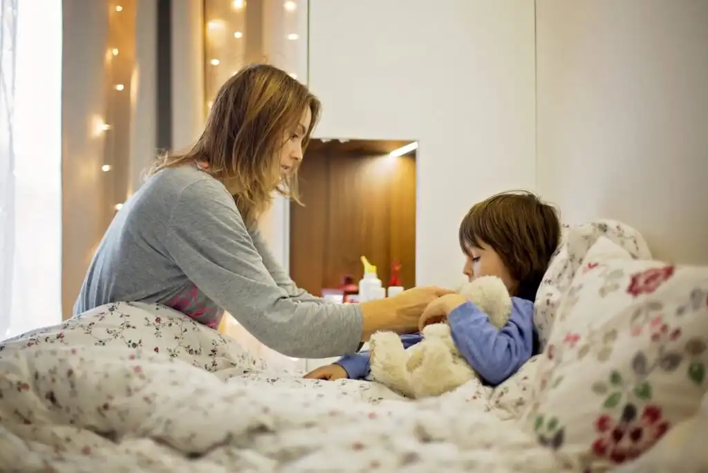 A woman in pajamas tends to a young child lying in bed with a teddy bear, in a softly lit bedroom with string lights in the background.