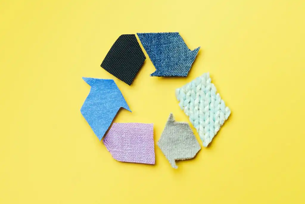 Recycling symbol made from fabric arrows of different colors and textures arranged in a circle on a yellow background.
