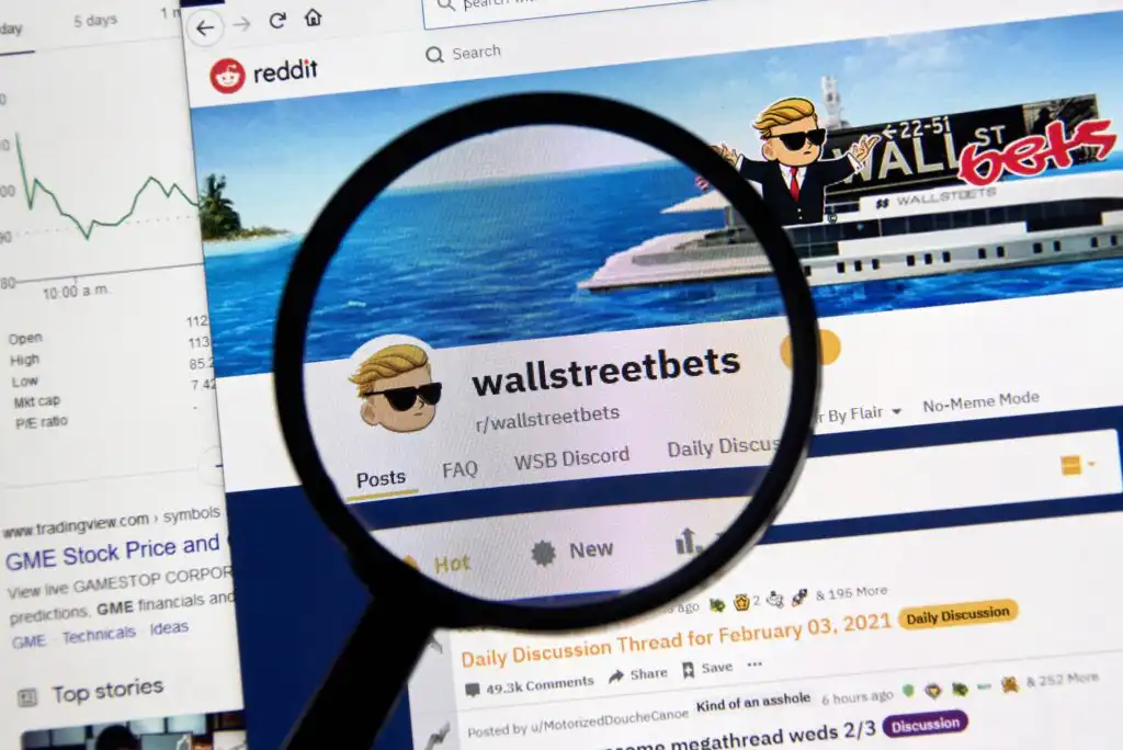 A magnifying glass focuses on the “wallstreetbets” subreddit page on Reddit, showing its profile banner with a cartoon character in sunglasses and a yacht. The webpage includes various tabs and discussion threads.