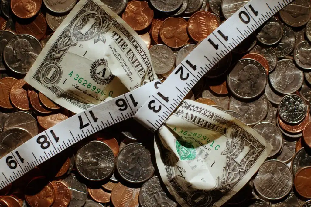 A crumpled one-dollar bill is cinched at the center by a white measuring tape, lying on a pile of assorted coins. The scene suggests financial constraint or budgeting.