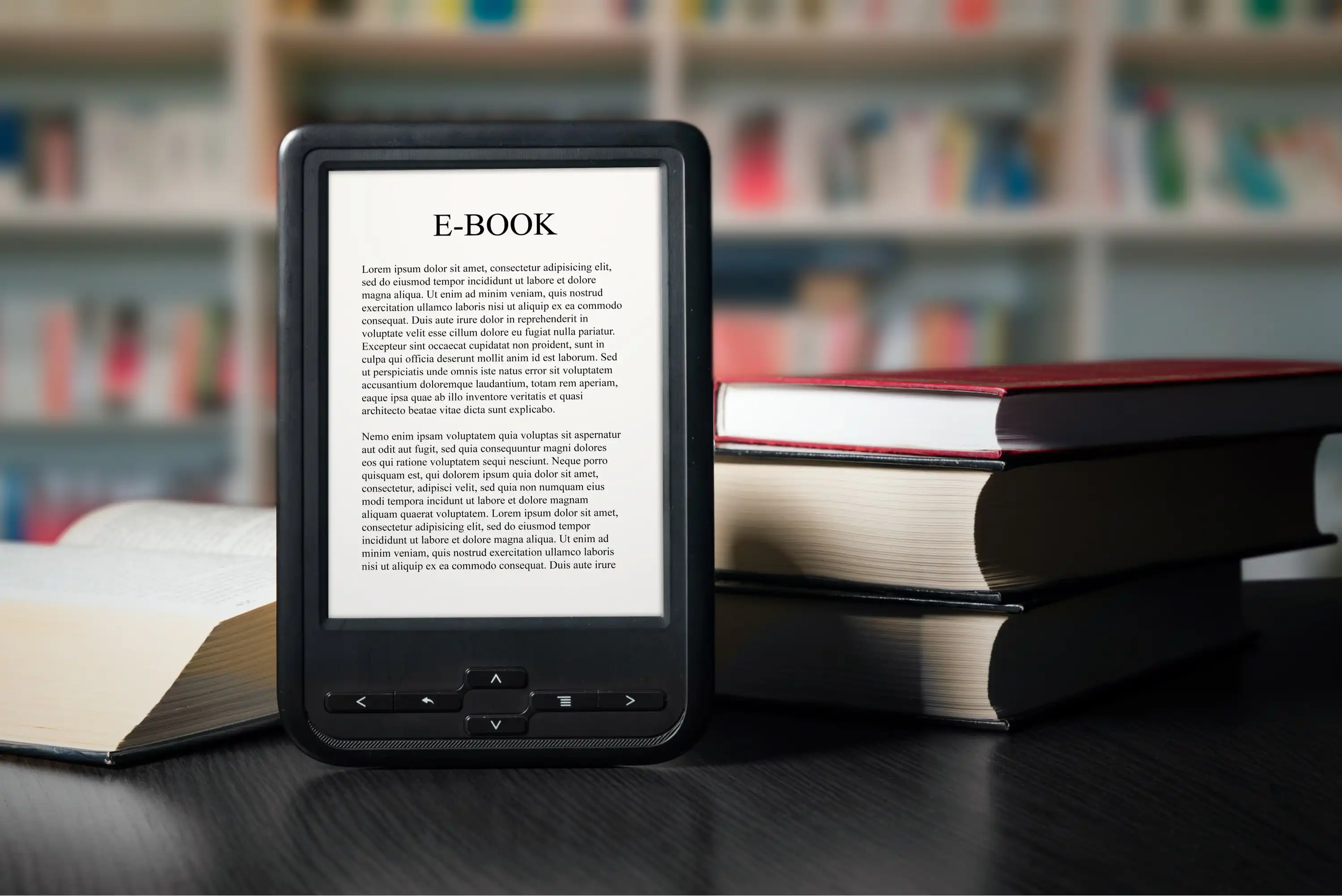An e-reader displaying an e-book is placed on a wooden table next to several stacked and open printed books, with a blurred bookshelf in the background.
