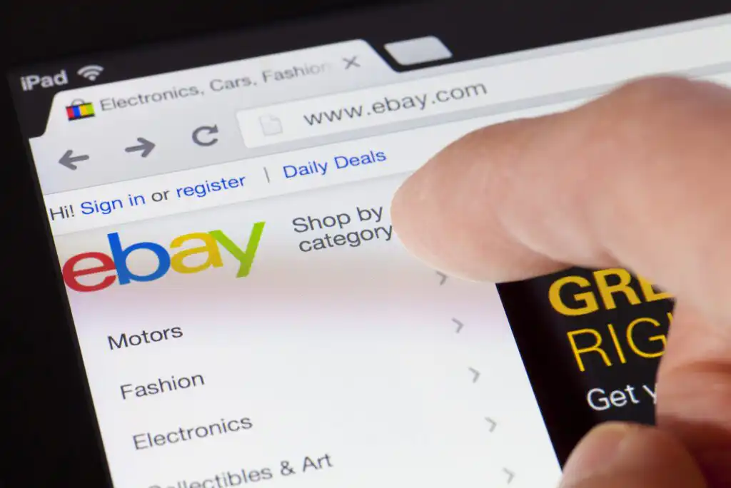 A close-up of a person using a tablet to browse eBay’s website, displaying the eBay logo, navigation menu with categories like Motors, Fashion, Electronics, and a finger tapping the screen.