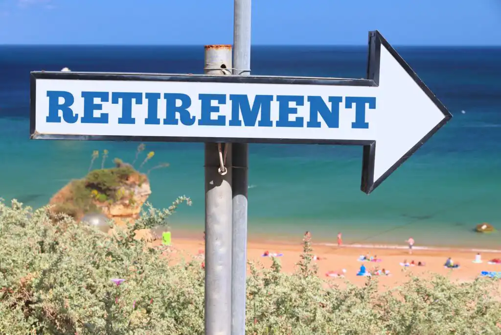 A large white arrow sign with the word RETIREMENT points right, positioned near a beach with people relaxing on the sand and greenery in the foreground. The ocean and blue sky are visible in the background.