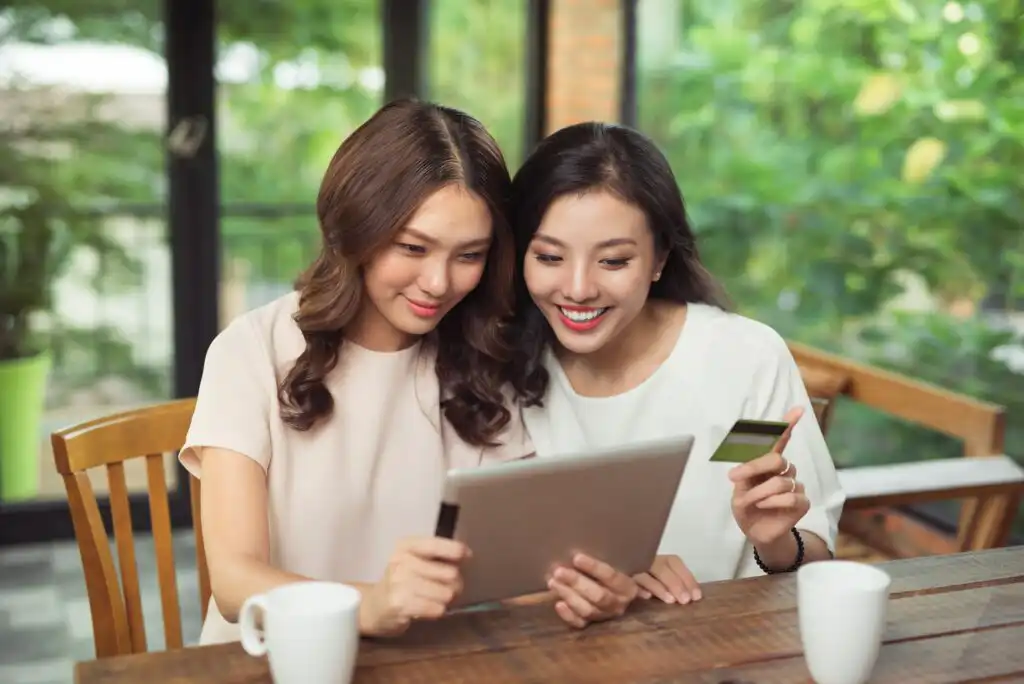 Two women sitting at a table with coffee mugs, smiling and looking at a tablet together; one woman is holding a credit card. They appear to be making an online purchase in a bright indoor setting with large windows.