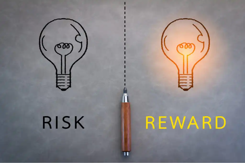 A pen divides a gray background; on the left is a black outline of a dim lightbulb labeled RISK, and on the right is a glowing lightbulb labeled REWARD in yellow letters.