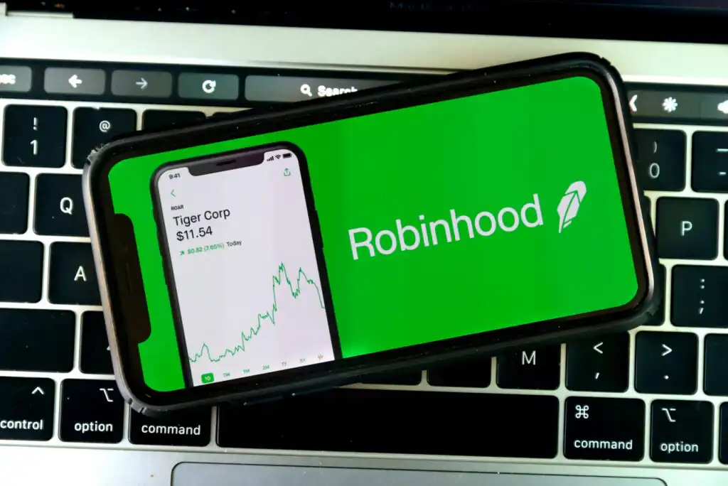 A smartphone displays the Robinhood app logo and a stock chart, resting on a laptop keyboard. The app screen shows a companys stock price and upward-trending graph.