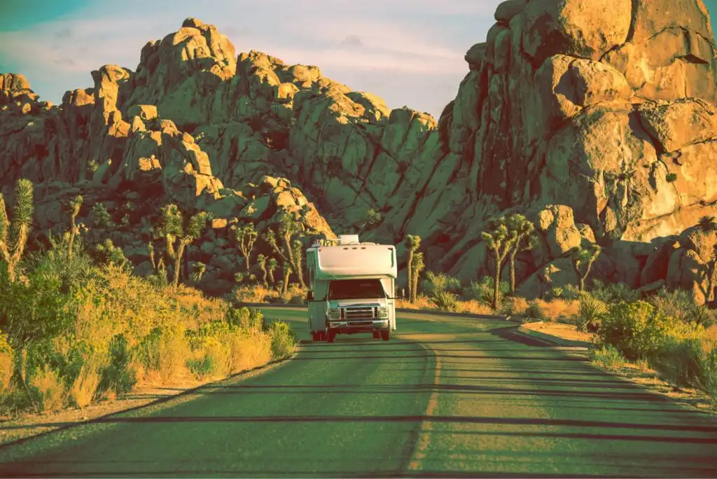 An RV drives down a paved road surrounded by desert vegetation and large rocky formations, under a partly cloudy sky during sunset.