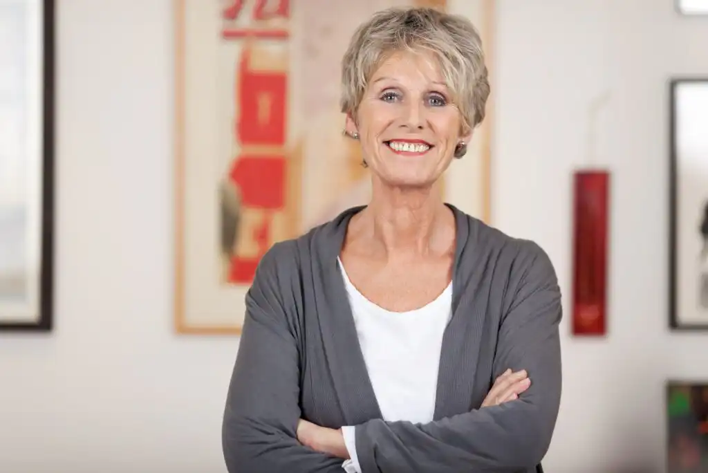 Smiling older woman with short gray hair stands with arms crossed, wearing a white shirt and gray cardigan, in a brightly lit room with artwork hanging on the walls in the background.
